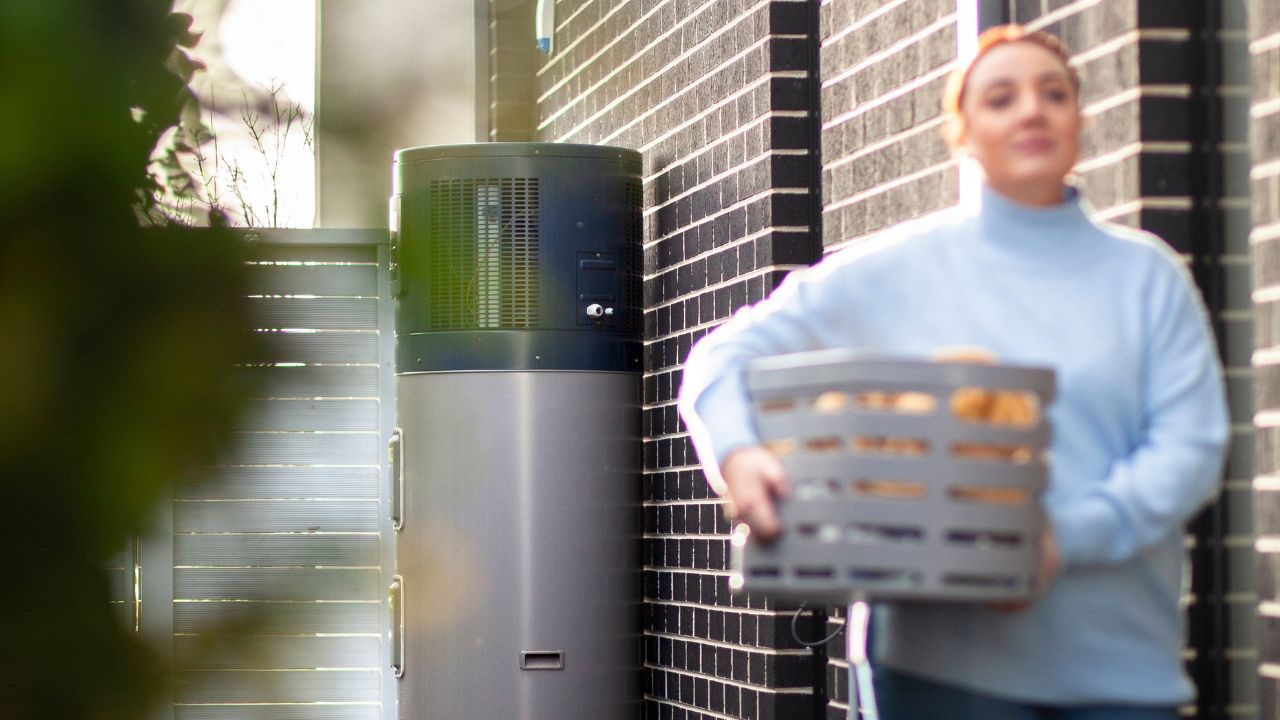 A woman walking outside holding a laundry basket with a hot water system in the background